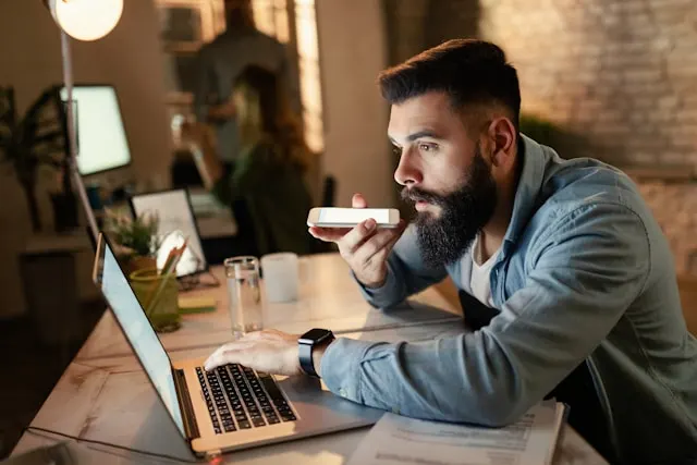A man uses voice-to-text on his phone while working on a laptop.